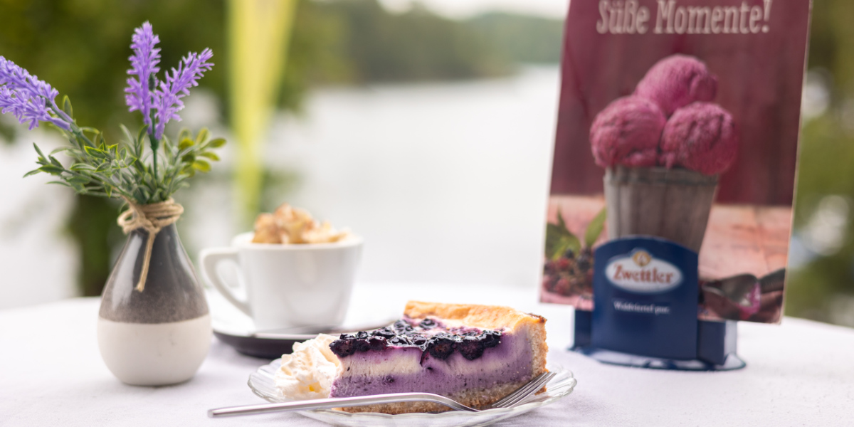 Ein stück Heidelbeerkuchen auf einem Teller mit einer Tasse Kaffee, draußen am See.